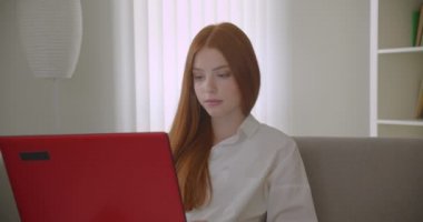 Dizüstü bilgisayar kullanarak genç güzel kızıl saçlı kız öğrencinin Closeup portre ve rahat bir dairede kanepede oturan kamera bakarak online eğitim