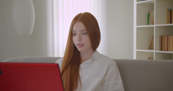 Retrato de close-up de jovem estudante ruiva bonita usando o laptop e estudando on-line olhando para a câmera feliz sentado no sofá em um apartamento acolhedor — Vídeo de Stock
