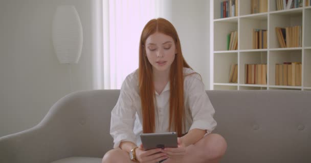 Närbild porträtt av unga ganska rödhårig kvinna använder tabletten sitter på soffan i en mysig lägenhet — Stockvideo