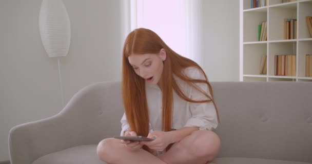 Gros plan portrait de jeune jolie rousse femelle parcourant la tablette assise sur le canapé dans un appartement confortable — Video