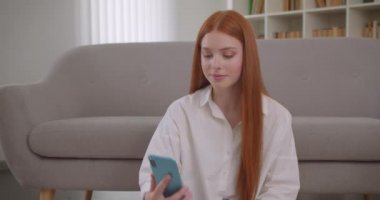 Telefonda bir görüntülü arama olan ve rahat bir dairede yerde oturan kamera önünde poz genç sevimli kızıl saçlı kadın Closeup portre