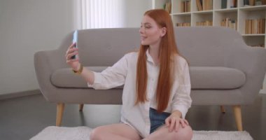 Rahat bir dairede yerde oturan telefonda selfie çeken genç güzel kızıl saçlı kadın Closeup portre