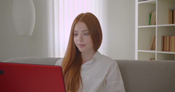 Крупним планом портрет молодої красивої рудої студентки, яка використовує ноутбук і вивчає онлайн, дивлячись на камеру весело сидячи на дивані в затишній квартирі — стокове відео