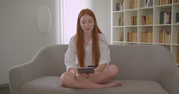 Портрет крупным планом молодой красивой рыжей женщины, рассылающей сообщения на планшете, сидящей на диване в уютной квартире — стоковое видео
