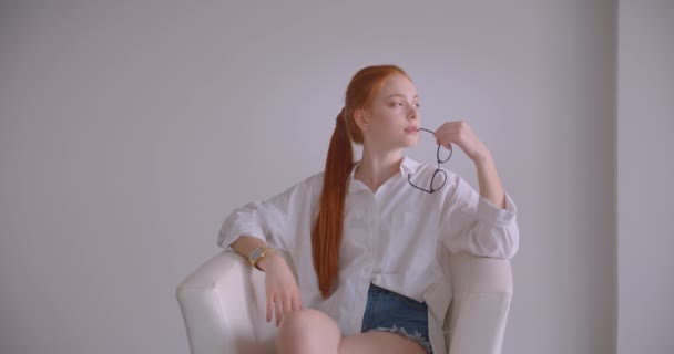 Primo piano ritratto di giovane bella rossa caucasica femmina guardando di lato tenendo gli occhiali con premura seduta sulla poltrona in una stanza bianca — Video Stock