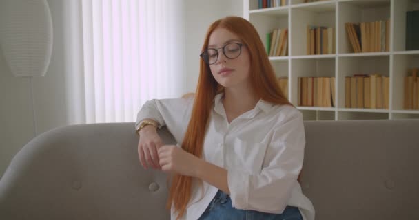 Zbliżenie portret młodego piękna Ruda Kobieta w okularach toczenia i patrząc na aparat uśmiechnięty wesoło siedzi na kanapie w przytulnym mieszkaniu w pomieszczeniu — Wideo stockowe