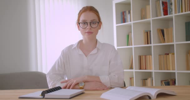 Retrato de cerca de una joven pelirroja pelirroja estudiante en gafas mirando a la cámara sonriendo felizmente sentada en el escritorio en un acogedor apartamento — Vídeos de Stock