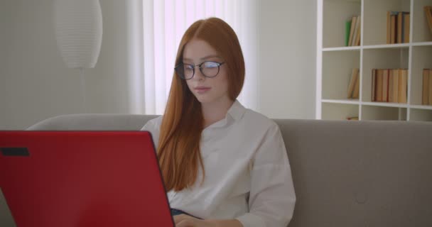 Primer plano retrato de joven pelirroja bonita estudiante en gafas usando el ordenador portátil y estudiando en línea mirando a la cámara felizmente sentado en el sofá en un acogedor apartamento — Vídeos de Stock