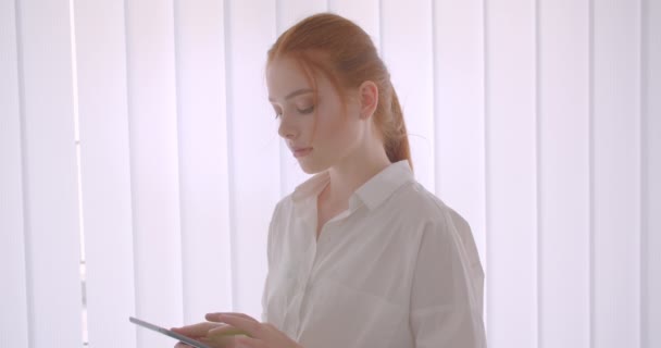 Крупный план портрета молодой красивой рыжей студентки, использующей планшет и показывающей зеленый экран перед камерой, стоящей в белой комнате — стоковое видео