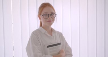 Beyaz odada duran kameraya bakan bir kitap tutan gözlük genç güzel kızıl saçlı kız öğrencinin Closeup portre