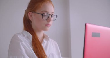 Gözlüklü genç beyaz kızıl saçlı genç iş kadınının yakın çekim portresi dizüstü bilgisayar kullanarak ofiste koltukta mutlu bir şekilde otururken gülümseyen kameraya bakıyor