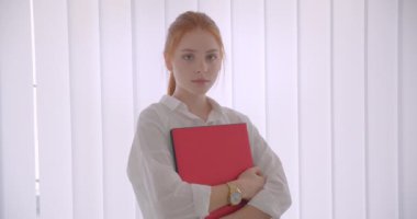 Bir ofiste duran kameraya bakarak dizüstü tutan genç güzel beyaz kızıl saçlı iş kadınınCloseup portre