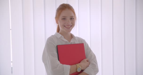 Primer plano retrato de joven bastante caucásica pelirroja mujer de negocios sosteniendo el portátil sonriendo felizmente mirando a la cámara de pie en una oficina — Vídeo de stock