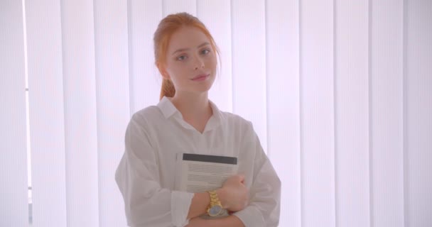Närbild porträtt av unga ganska rödhårig kvinnlig student som innehar en bok som tittar på kameran stående i vita rummet — Stockvideo