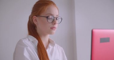 Gözlüklü genç sevimli beyaz kızıl saçlı iş kadınının yakın çekim portresi dizüstü bilgisayar kullanarak ofiste koltukta mutlu bir şekilde otururken gülümseyen kameraya bakıyor