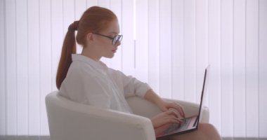 Ofiste koltukta oturan dizüstü bilgisayar üzerinde çalışan gözlükgenç güzel beyaz kızıl başlı iş kadının Closeup yan görünüm portresi