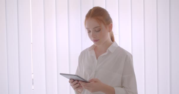 Крупним планом портрет молодої красивої рудої студентки, яка використовує планшет, дивлячись на камеру, посміхаючись щасливо стоячи в білій кімнаті — стокове відео