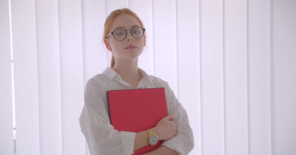 Κοντινό πλάνο της νεαρής όμορφη καυκάσια κοκκινομάλλα επιχειρηματίας σε γυαλιά κρατώντας το φορητό υπολογιστή κοιτάζοντας την κάμερα στέκεται σε ένα γραφείο — Αρχείο Βίντεο