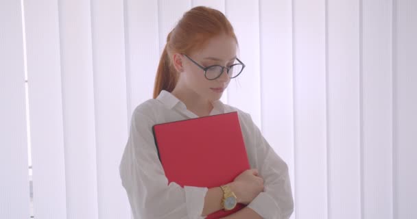 Primo piano ritratto di giovane bella donna d'affari rossa caucasica in occhiali che tiene il computer portatile guardando la fotocamera sorridente felicemente in piedi in una stanza bianca — Video Stock