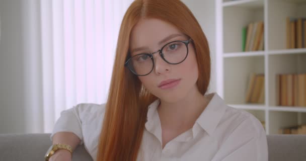 카메라를보고 실내 아늑한 아파트의 소파에 앉아 포즈를 취하는 안경을 쓴 젊은 빨간 머리 여성의 클로즈업 초상화 — 비디오