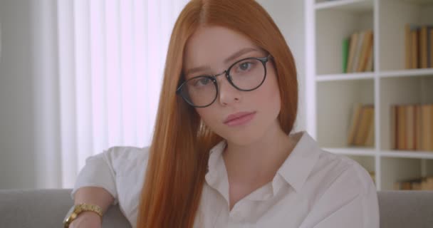 Closeup portrait of young redhead female in glasses looking at camera and posing sitting on the couch in the cozy apartment indoors — Stock Video