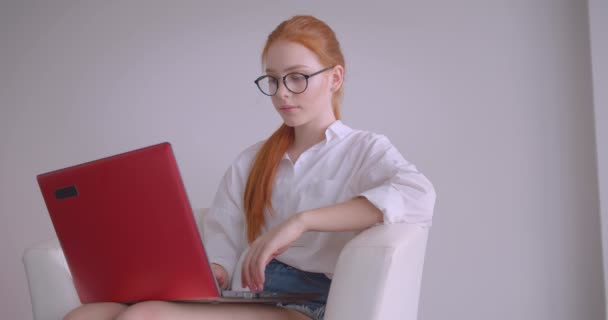 戴着眼镜的年轻漂亮的白种红发女商人的特写肖像，用笔记本电脑看着相机，微笑着愉快地坐在白色房间的扶手椅上 — 图库视频影像