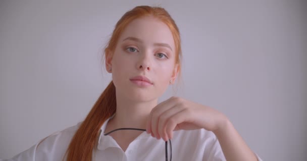 Närbild porträtt av unga söta kaukasiska rödhårig kvinna tittar på kameran håller hennes glasögon med omtänksamhet sitter i fåtöljen i ett vitt rum — Stockvideo