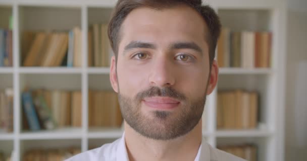 Nahaufnahme Porträt eines erwachsenen gutaussehenden kaukasischen Mannes, der glücklich in die Kamera lächelt, in der Bibliothek mit Bücherregalen im Hintergrund drinnen — Stockvideo