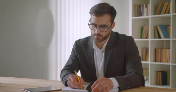 Крупним планом портрет дорослого гарного кавказького бізнесмена в окулярах, використовуючи планшет, приймаючи нотатки в офісі в приміщенні — стокове відео