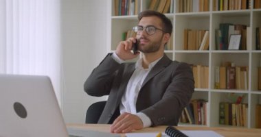 Arka planda kitaplıklar ile ofiste kapalı laptop önünde oturan bir telefon görüşmesi sahip gözlük yetişkin yakışıklı beyaz işadamı bir portre