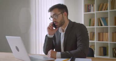 Yetişkin yakışıklı sakallı beyaz işadamı dizüstü bilgisayar kullanarak gözlük ve ofis kapalı bir telefon görüşmesi olan portre