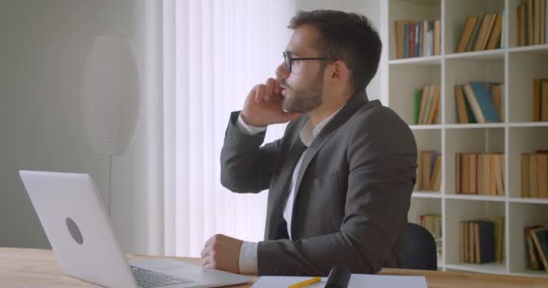 Closeup portrait of adult handsome bearded caucasian businessman in glasses using the laptop and having a phone call in the office indoors with bookshelves on the background — Stock Video