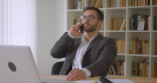 Closeup portrait of adult handsome caucasian businessman in glasses having a phone call sitting in front of the laptop in the office indoors with bookshelves on the background — Stock Video