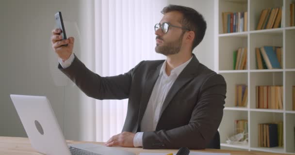 Κοντινό πλάνο του ενήλικα όμορφο μούσι Καυκάσιος επιχειρηματίας σε γυαλιά παίρνοντας selfie των στο τηλέφωνο κάθεται μπροστά από το φορητό υπολογιστή στο γραφείο σε εσωτερικούς χώρους με ράφια στο παρασκήνιο — Αρχείο Βίντεο