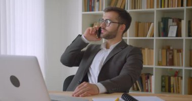 Arka planda kitaplıklar ile ofiste kapalı laptop önünde oturan bir telefon görüşmesi sahip gözlük yetişkin yakışıklı sakallı Kafkas işadamı portre