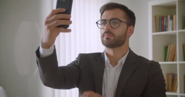 Arka planda kitaplıklar ile ofiste kapalı telefonda selfie 'ler alarak gözlük yetişkin yakışıklı sakallı beyaz işadamı portre