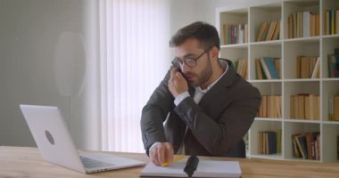Dizüstü bilgisayar kullanarak gözlüklü yetişkin yakışıklı beyaz işadamı portre ve ofis kapalı notlar alarak bir telefon görüşmesi sahip