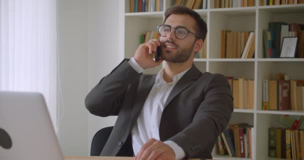 Portrét dospělého úspěšného podnikatele v brýlích s telefonem, který sedí před notebookem v kanceláři v interiéru s knižními regály na pozadí — Stock video