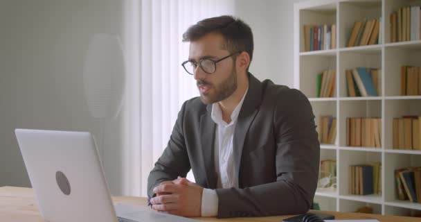 Nahaufnahme Porträt eines erwachsenen gutaussehenden bärtigen kaukasischen Geschäftsmannes mit Brille, der einen Videoanruf auf dem Laptop im Büro hat, drinnen mit Bücherregalen im Hintergrund — Stockvideo