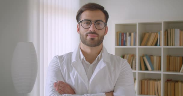 Retrato de close-up de adulto bonito branco médico masculino em casaco branco e óculos olhando para a câmera na biblioteca com estantes no fundo dentro de casa — Vídeo de Stock