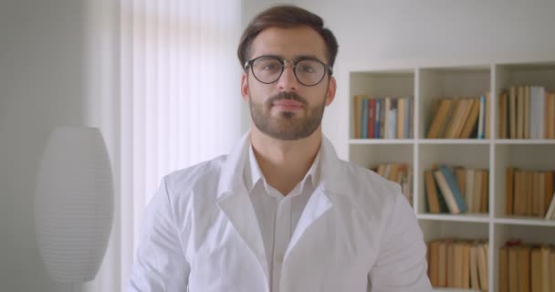 Primo piano ritratto di adulto bel medico maschio caucasico in occhiali guardando la fotocamera sorridente felicemente in biblioteca con librerie sullo sfondo all'interno — Video Stock