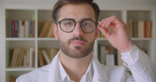 Nahaufnahme Porträt eines erwachsenen gutaussehenden kaukasischen Geschäftsmannes, der in die Kamera blickt und seine Brille in der Bibliothek mit Bücherregalen im Hintergrund im Inneren befestigt — Stockvideo