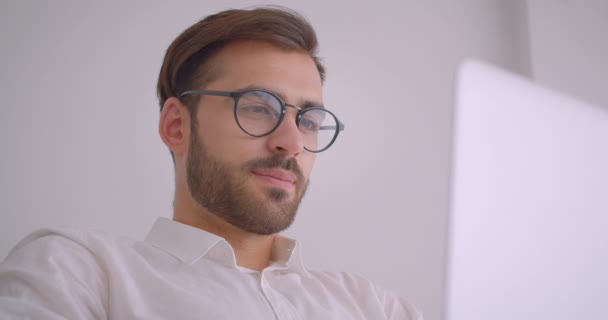 Крупный план портрет красивого бородатого кавказского бизнесмена в очках, использующего ноутбук, смотрящего на камеру, счастливо улыбающегося сидя в кресле в белом кабинете в помещении — стоковое видео