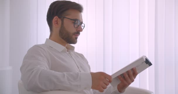 Closeup pohled na portrét dospělého krasavce v brýlích, který si přečetl knihu, sedící v křesle v bílé kanceláři — Stock video