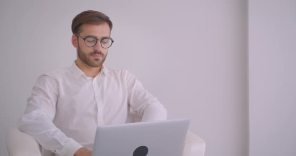 Primo piano ritratto di adulto bello uomo d'affari caucasico di successo in occhiali usando il computer portatile guardando la macchina fotografica sorridente allegramente seduto nella poltrona nell'ufficio bianco all'interno — Video Stock