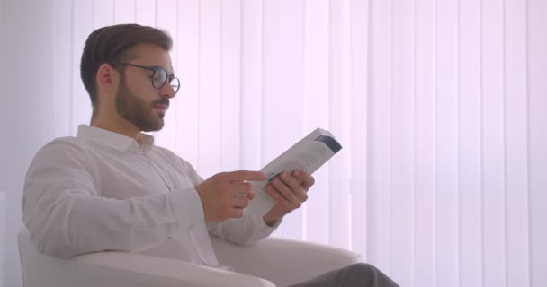 Closeup side view portrait of adult handsome bearded caucasian businessman in glasses reading a book sitting in the armchair in the white office indoors — Stock Video