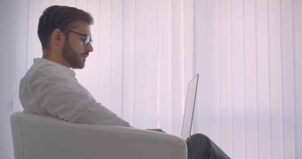 Primer plano vista lateral retrato de adulto guapo barbudo hombre de negocios caucásico en gafas usando el portátil sentado en el sillón en la oficina blanca en el interior — Vídeos de Stock