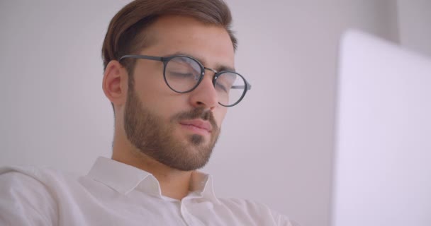 Primer plano retrato de guapo barbudo hombre de negocios caucásico en gafas usando el portátil sentado en el sillón en la oficina blanca en el interior — Vídeos de Stock