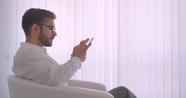 Primer plano vista lateral retrato de adulto guapo barbudo hombre de negocios caucásico en gafas usando la tableta sentada en el sillón en la oficina blanca en el interior — Vídeos de Stock