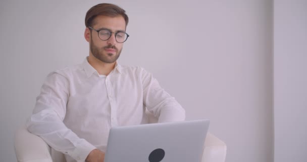 Close-up portret van volwassen knappe bebaarde Kaukasische zakenman in glazen met behulp van de laptop kijken naar camera glimlachend vrolijk zittend in de fauteuil in het witte kantoor binnenshuis — Stockvideo